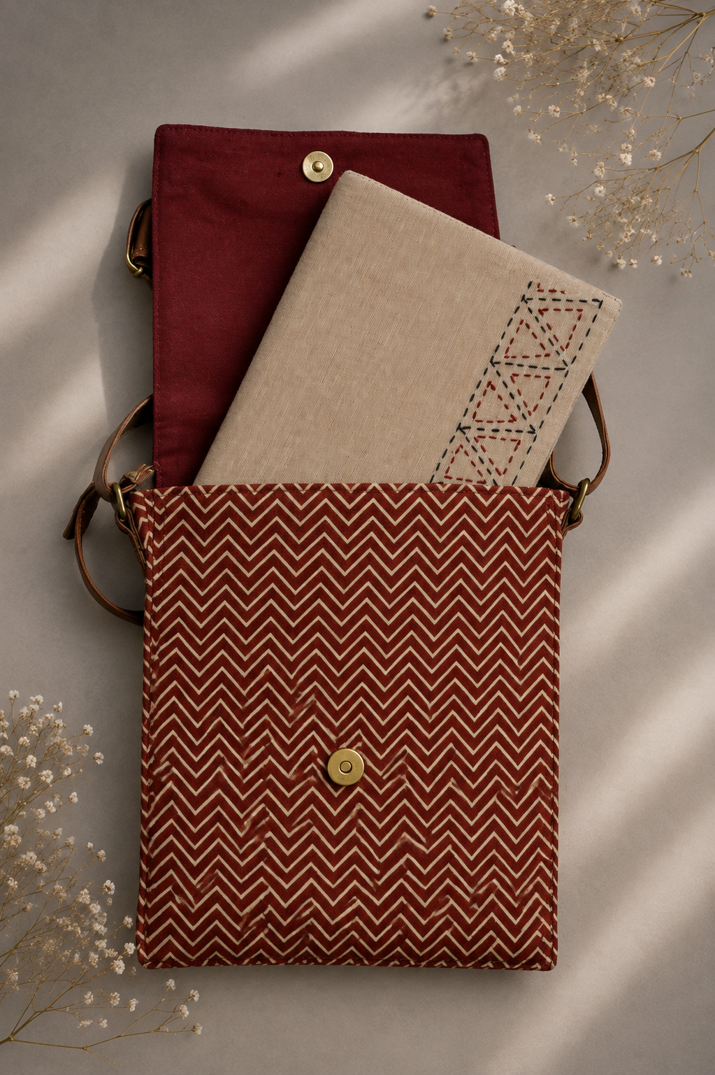 A structured fabric sling cum document bag with a rust-red and white chevron patterned front flap, shown open with a beige embroidered notebook or folder tucked inside. The bag has a dark red interior lining, brown leather shoulder straps, and a gold-tone snap button closure.